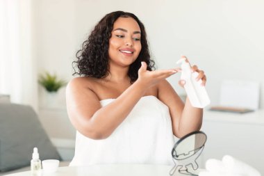 Vücut geliştirici. Güzel, siyah, iri yarı bir kadın elinde nemlendirici losyon, yatak odasında iç tarafta oturuyor ve evde cilt bakımı yapıyor.