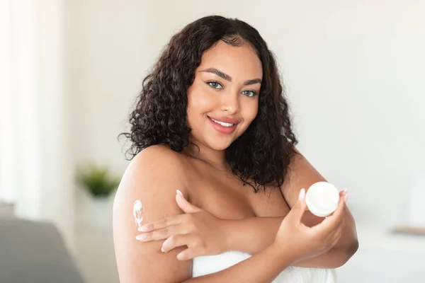 新鮮な肌 保湿と体のための栄養 正のアフリカ系アメリカ人プラスサイズの女性が家で朝の肌のためのクリームを使用してタオルに包まれ 空のスペース — ストック写真