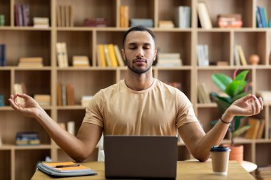 İş yeri stres yönetimi. Sakin bir Arap, laptopun önünde gözleri kapalı meditasyon yapıyor, evde masa başında oturuyor. Genç erkek serbest çalışan, kendini huzurlu ve dengeli hissediyor.
