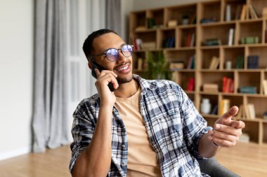 Mutlu Arap adam akıllı telefondan konuşuyor ve oturma odasındaki koltukta dinlenirken gülümsüyor. Genç adam hoş bir telefon görüşmesi yapıyor, evde iletişim kuruyor.