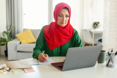 Laptop kullanan, bütçeyi hesaplayan, oturma odasındaki masada oturan tesettürlü müslüman bir kadın. Modern İslami kadın serbest bilgisayar üzerinde çalışıyor.