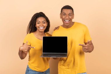 Güzel web sitesi konsepti. Mutlu siyah eşler boş ekranlı dizüstü bilgisayarı işaret ediyor, olumlu adam ve kadın online reklam için fotokopi alanı gösteriyor, şeftali rengi stüdyo arka planının başında duruyor,