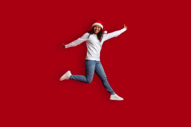 Mutlu Noeller ve mutlu yıllar 2023. Duygusal, kıvırcık, siyah bir kadın Noel Baba şapkası takıyor kırmızı stüdyo arka planında zıplıyor, eğleniyor.