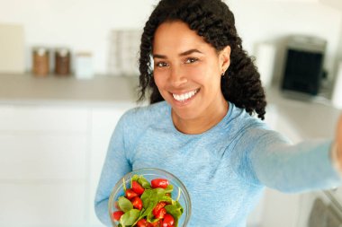 Afro-Amerikan formda kadın elinde taze sebze salatasıyla kaseyle mutfakta selfie çekiyor ve sağlıklı beslenmeyi öneriyor. Dengeli yemek yiyen heyecanlı kadın portresi