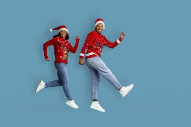 Kırmızı Xmas süveter ve Noel Baba şapkalı komik Afro-Amerikan erkek ve kadın mavi stüdyo arka planında zıplıyorlar, kış tatillerinin tadını çıkarıyorlar, yeni yıl 2023, kopyalama alanı, tam boy