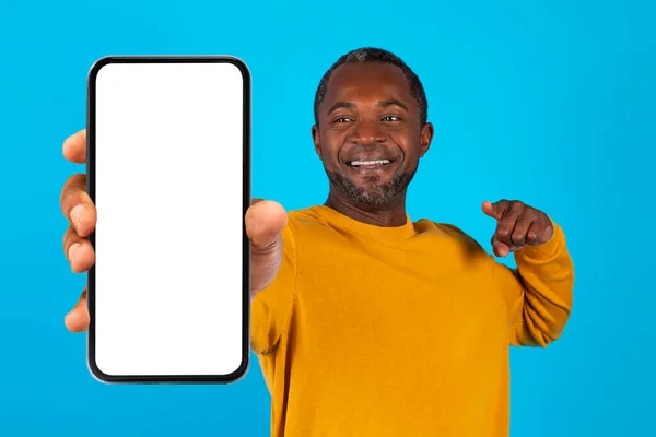 英俊潇洒的中年非洲裔美国男人指着带有白屏的全新手机 推荐最新的移动应用程序 在蓝色工作室背景下被隔离 — 图库照片