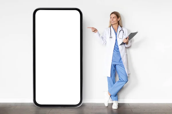 站在医院白墙边的大空白智能手机屏幕上 穿着制服指尖手指的女医生微笑 女医生示范医疗广告的复制空间 — 图库照片