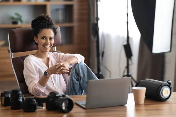 stock image Happy beautiful brunette young woman influencer photographer working at office, sitting at workdesk in front of laptop with camera and various lenses on, smiling at camera, copy space