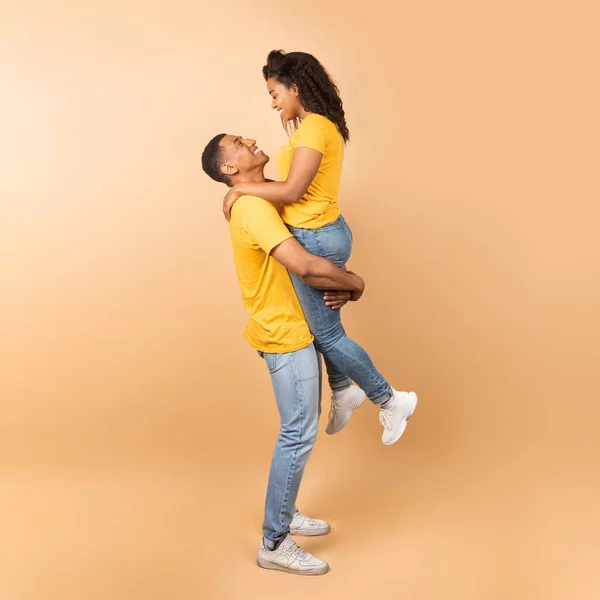 喜欢的黑人男人抬起他的女朋友 抱着她在桃子工作室的背景 侧面看 全身拍摄 一对浪漫的情侣彼此表达爱意 — 图库照片