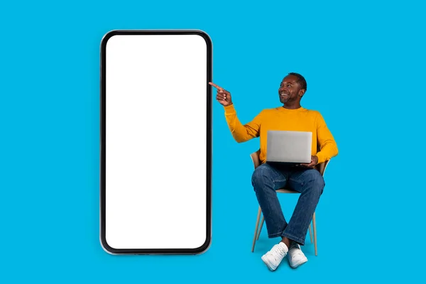 快乐的中年黑人男子 穿着休闲的自由职业者 坐在蓝色工作室的椅子上 使用现代电脑 指着有白色空屏幕的大手机 模仿着 商业移动应用程序 — 图库照片
