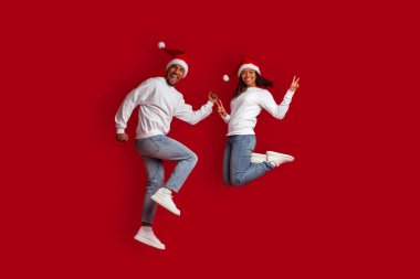 Noel Baba şapkası takan, gülümseyen ve el kol hareketi yapan, yeni yıl 2023 'ü birlikte kutlayan, kırmızı stüdyo arka planında izole edilmiş, genç ve güzel siyah bir çift.