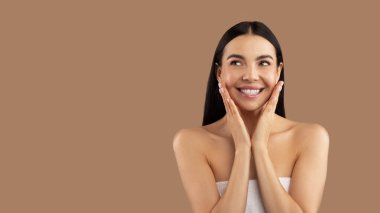 Yüzünde yarı çıplak, binlerce yıllık güzel esmer kadın, yanaklarına dokunduktan sonra havluya sarılmış, fotokopi odasına bakıyor, organik cilt ürünlerinin tadını çıkarıyor, bej renkli stüdyo arka planında izole edilmiş.