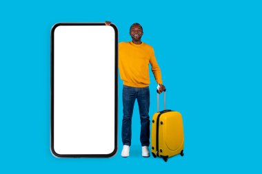 Heyecanlı Afro-Amerikan orta yaşlı adam tek başına seyahat ediyor, büyük bir telefonla poz veriyor mavi ekranda izole edilmiş, sarı bavul taşıyor, stüdyo fotoğrafı çekiyor, mobil uygulama kullanıyor.