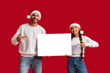 Noel Baba şapkalı mutlu bir çift ellerinde boş bir plaketle kırmızı arkaplanı işaret ediyor neşeli bir Orta Doğulu erkek ve kadın Noel listesi ve reklam için kopya alanı gösteriyor.
