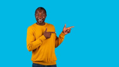 Havalı, mutlu, orta yaşlı, sarı jestli siyah adam fotokopi alanını gösteriyor ve gülümsüyor. Güzel bir teklif veya anlaşma öneriyor. Promosyon, mavi stüdyo geçmişi, panorama.