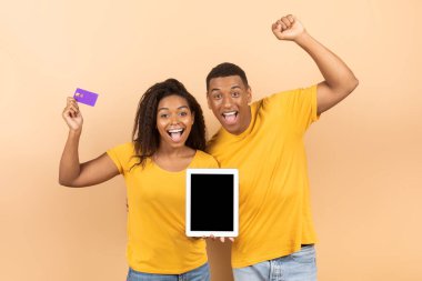 Kredi kartı tutan ve şeftali stüdyosunun arka planında boş ekranlı dijital tableti gösteren mutlu siyah eşler, yeni online alışveriş uygulaması tasarımı için model