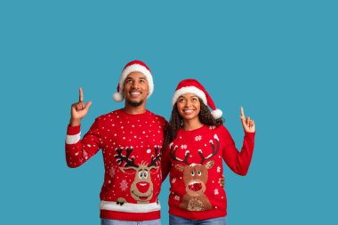 Mutlu çekici siyah erkek ve kadın birbirini seven çiftler, reklam için fotokopi alanına işaret ediyorlar, harika xmas teklifleri gösteriyorlar, Noel Baba şapkaları giyiyorlar, mavi stüdyo arka planında izole edilmiş.