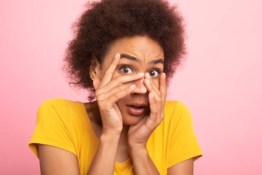 Bin yıllık korkak Afro-Amerikan kıvırcık kadın öğrenci sarı tişörtlü yüzünü ellerle örttü ve korktu, pembe arka planda, stüdyoda izole edildi. İnsani duygular, yüz ifadesi ve kötü haberler.
