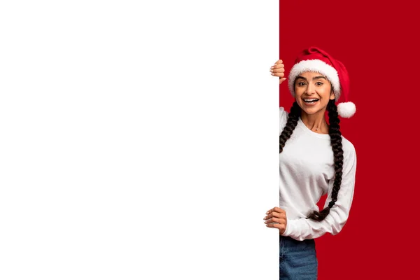 サンタハットに立つ美しいアラブの女性ブランクホワイト広告ボードの近くに立つ 陽気な若い中東の女性クリスマスマストの願いリストや販売の提供のためのコピースペースを示す モックアップ — ストック写真