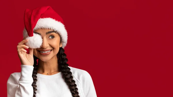 Xmas サンタハットでポンポンで目を覆い 赤い背景の上にコピースペースを脇に見て 幸せな興奮した中東の女性クリスマスオファー パノラマを楽しむ — ストック写真