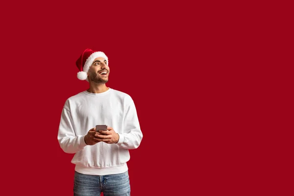 快乐的阿拉伯男子 头戴圣诞帽 手持智能手机 遥望远方 快乐的中东年轻人 手拿手机 与红色背景隔离 复制空间 — 图库照片