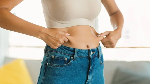 Young woman in jeans pinching fat of her abdomen zone, unrecognizable lady ready for slimming or liposuction treatment, posing at home, panorama, cropped