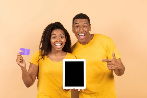 Happy black man pointing at empty tablet screen, his girlfriend holding credit card on peach studio background, mockup for design. Excited couple promoting online shopping