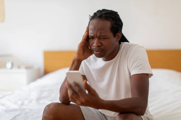 悪い知らせだ ストレスを受けた黒人男性がベッドに座ってスマートフォンの画面を見て 心配している若いアフリカ系アメリカ人男性が頭に触れながら不快なメッセージを読み取り コピースペースでショットを閉じます — ストック写真