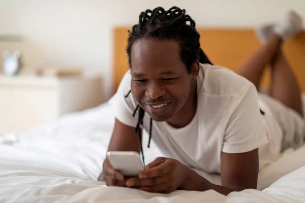友人との陽気な若い黒人男性メッセージングや自宅でベッドに横たわっている間 スマートフォンでインターネットを閲覧 ベッドルームでリラックスしながら携帯電話を使用して幸せなアフリカ系アメリカ人の男 — ストック写真