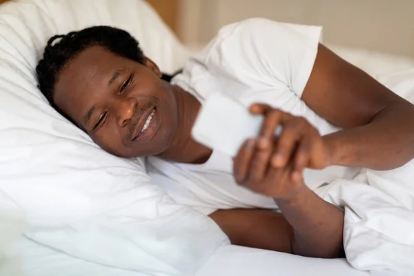 目覚めの後にベッドに横たわっている間 スマートフォンで幸せな黒人男性メッセージング 笑顔若いアフリカ系アメリカ人の男は自宅でベッドルームで休んでいる間 携帯電話にテキストやソーシャルメディアを閲覧します — ストック写真