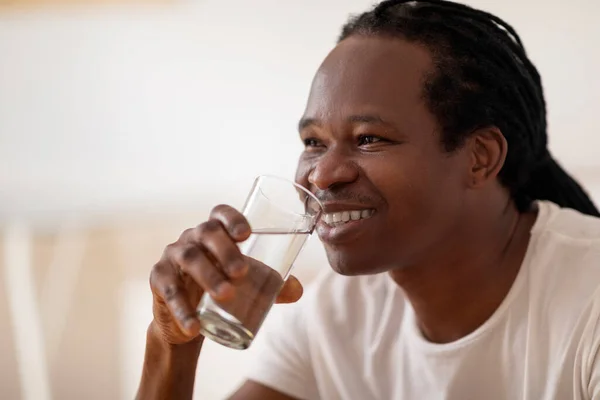 ウェルネスコンセプト 家でリラックスしながらガラスから水を飲むハンサムな黒人男性の閉鎖ショット 幸せな若いアフリカ系アメリカ人男性はさわやかなミネラルドリンクを楽しんで フリースペース — ストック写真