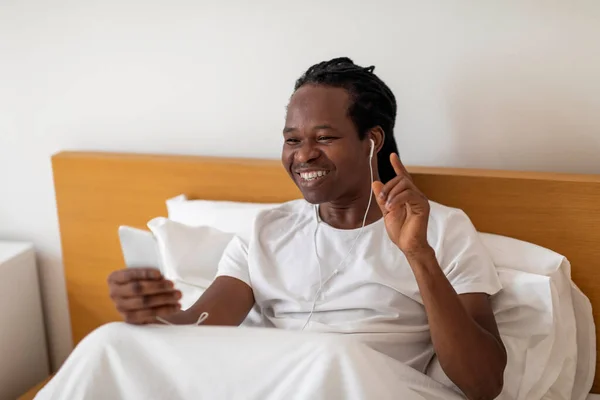 スマートフォンやイヤフォンでベッドでリラックスして陽気な黒人男性 自宅でお気に入りの音楽を聴いて幸せな若いアフリカ系アメリカ人の男 ベッドルームで休んでいる間にオンラインプレイリストを楽しみます — ストック写真