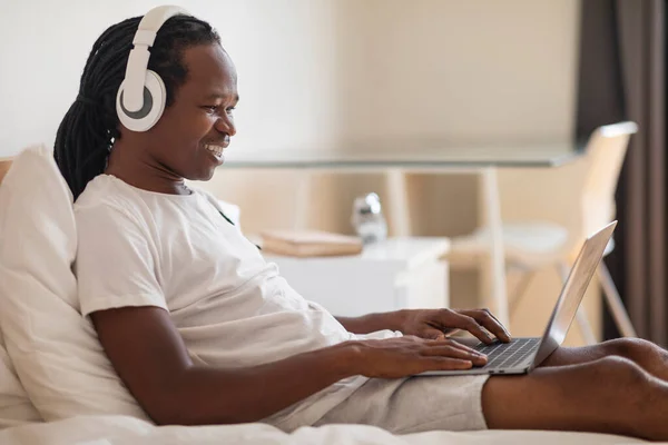 Lächelnder Schwarzer Mann Drahtlosen Kopfhörern Der Bett Auf Dem Laptop — Stockfoto