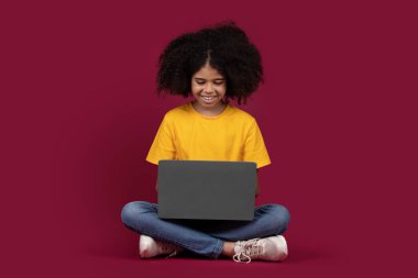 Gündelik kıyafet okulunda parlak saçlı neşeli siyah kız modern dizüstü bilgisayar kullanıyor, renkli bordo renkli stüdyo arka planında izole edilmiş, kopyalama alanı kullanıyor. Çocuklar ve alet bağımlılığı