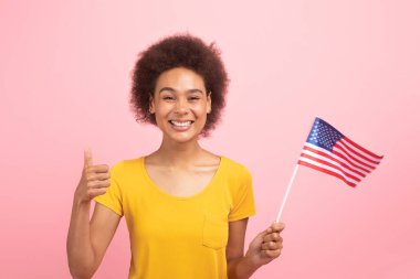 Sarı tişörtlü mutlu Afro-Amerikan kıvırcık kadın öğrenci başparmağını kaldırdı, ABD bayrağı, pembe arka planda, stüdyoda soyutlandı. El işareti, İngilizce dersi, tavsiye, değişim eğitimi ve eğitim