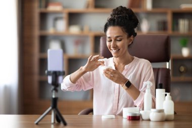 Neşeli, dost canlısı, çekici esmer kadın güzellik blogcusu yüz bakımını öneriyor, evden video veya yayın yapıyor, çeşitli kozmetik ürünlerini gösteriyor, cep telefonu kullanıyor.