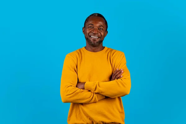 陽気な自信を持ってハンサムな中年アフリカ系アメリカ人男性で黄色笑顔でカメラ ポーズだけで青のスタジオの背景に 保持腕クロス胸 コピースペース — ストック写真