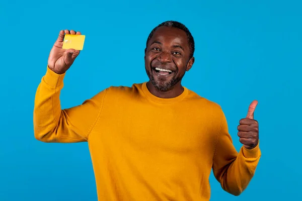 快乐的 情绪化的 英俊的非洲裔美国成年男子 身穿黄色衣服 在网上推荐申请信用卡 面带微笑 在蓝色的工作室背景下与世隔绝 — 图库照片