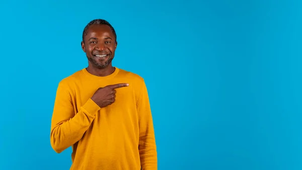 クール陽気な魅力的なアフリカ系アメリカ人中年男で黄色指していますコピースペースのために広告と笑顔以上青スタジオ背景 パノラマ モックアップのためにテキスト — ストック写真