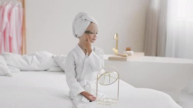 Evde spa oynayan, guaj silindirli cilt masajı yapan, kafasında havluyla bornozla evde oturan, ağır çekimde, boş bir alanda oturan tatlı küçük bir kız.