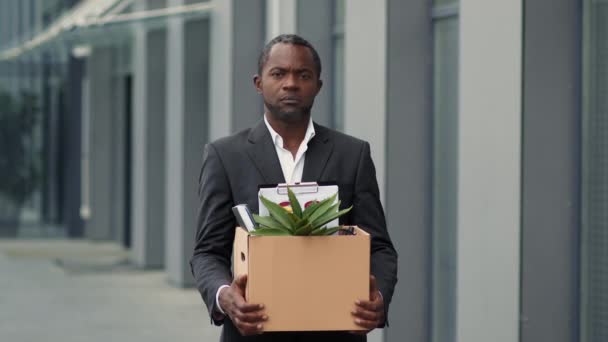 企業差別問題 解雇後にオフィスビルの近くに立つアフリカ系アメリカ人男性従業員の動揺の屋外の肖像画 個人的な持ち物と箱を運ぶ スローモーション フリースペース — ストック動画