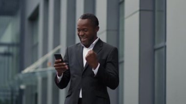 Mutlu orta yaşlı Afro-Amerikan iş adamı akıllı telefondan e-posta veya mesaj okuyor başarılı bir iş birliğinin tadını çıkarıyor ofis binasının yanında duruyor, yavaş çekim, boş alan