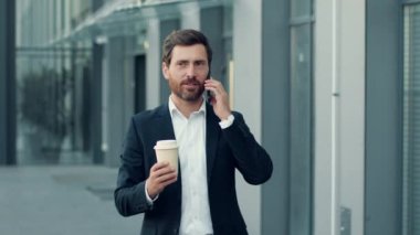 Kendine güvenen orta yaşlı işadamı iş arkadaşlarıyla telefonda konuşuyor, ofis binasının yakınındaki kahvelerle yürüyor, çekimi takip ediyor, yavaş çekim, serbest alan