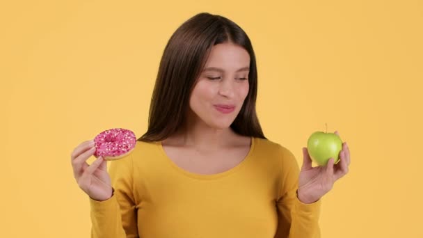 健康和天然的糖果 年轻的情绪孕妇在甜甜圈和有机苹果之间做出选择 吃新鲜水果 有黄色的工作室背景 动作缓慢 — 图库视频影像