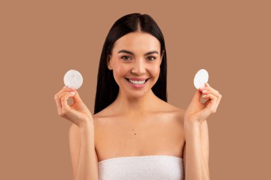 Makyaj makinesi, toner, temizleyici süt. Güzel, yarı çıplak esmer, uzun saçlı, banyo havlusuna sarılı, elinde pamuk pedlerle gülümseyen, bej renkli stüdyo arka planında soyutlanmış yetişkin bir kadın.