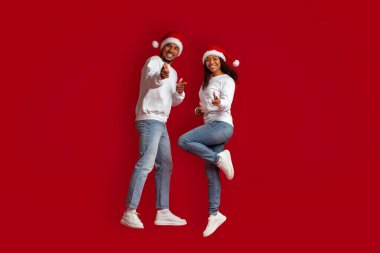 Noel Baba şapkası takmış neşeli, genç siyah bir çift kırmızı stüdyo arka planında kameraya doğru zıplıyor, Noel ve yeni yıl 2023 'ü birlikte kutluyor, kopyalama alanı, tam boy
