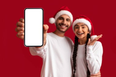 Xmas Teklif. Noel Baba Şapkalı Genç Arap Çift Büyük Beyaz Ekranlı Boş Akıllı Telefon Gösterileri Mutlu Orta Doğulu Erkek ve Kadın Yeni Uygulama veya Web Sitesi Tavsiye Ediyor