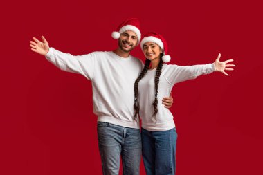 İyi tatiller. Noel Baba şapkaları takan ve kameraya gülümseyen neşeli Orta Doğu eşleri Noel tatilleriyle selamlayan, kırmızı arka planda duran neşeli genç Arap çift.