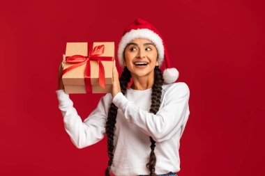 Tatil havası. Heyecanlı Arap Kadın, Noel Baba Şapka Giyen Paketli Güzel Mutlu Orta Doğulu Kadın Xmas 'ı Sarsıyor Kırmızı Stüdyo Arkaplanı Üzerinde Yalnız Poz Veriyor, Boşluğu Kopyala