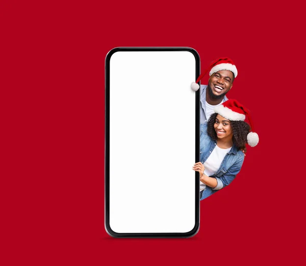 Happy black millennial lady and guy in Santa Claus hats peek out from big smartphone with empty screen isolated on red background, studio. New Year and Christmas celebrations, great offer, ad and app
